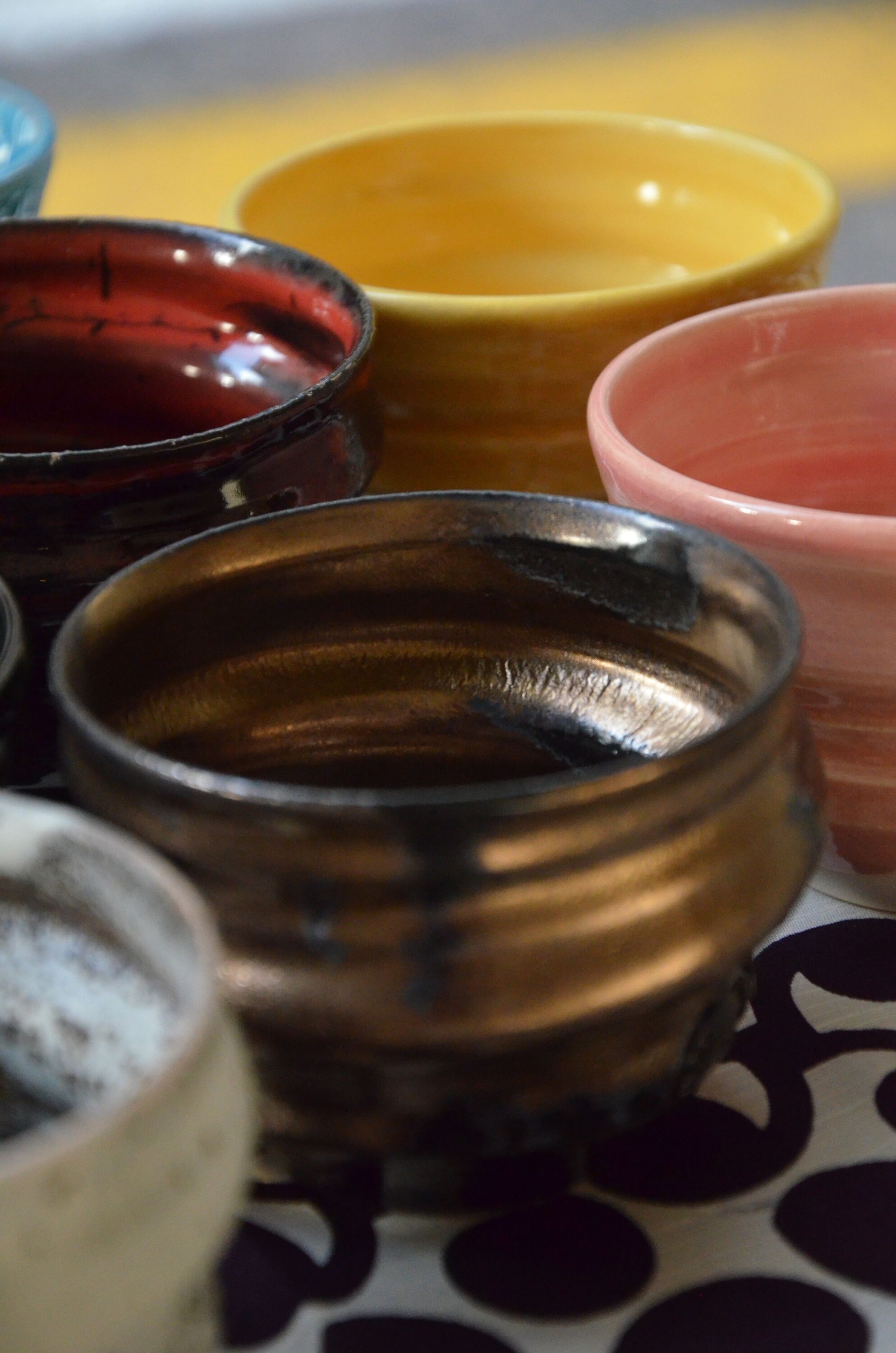 A close-up view of a collection of Ninshu handmade ceramic cups, showcasing their unique textures and colors. The cups are arranged closely together, with one displaying a dark, glossy brown glaze that reflects light, another featuring a deep red with a slightly cracked surface, and others in soft pink and bright yellow. The background features a patterned cloth with black and white polka dots, providing a striking contrast to the rich, earthy tones and smooth surfaces of the artisanal ceramics.