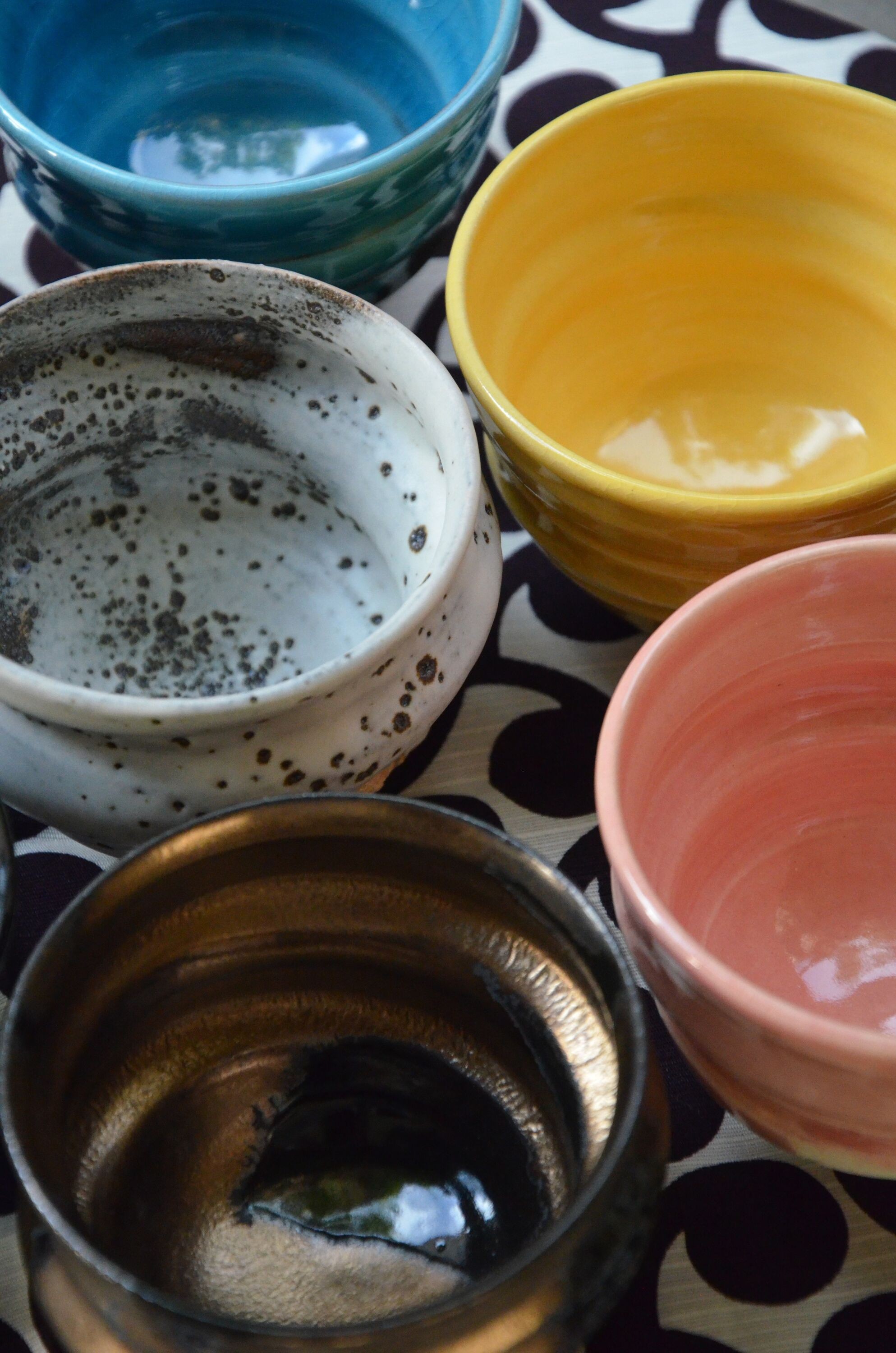 A collection of five Ninshu ceramic cups, photographed from above to show their interiors. The cups display a variety of colors: one white with dark spots and signs of use, one grey with streaks, one dark blue, one light pink, and one bright yellow. The cloth underneath has a black polka-dot pattern on a white background, contrasting with the vibrant colors of the ceramics.