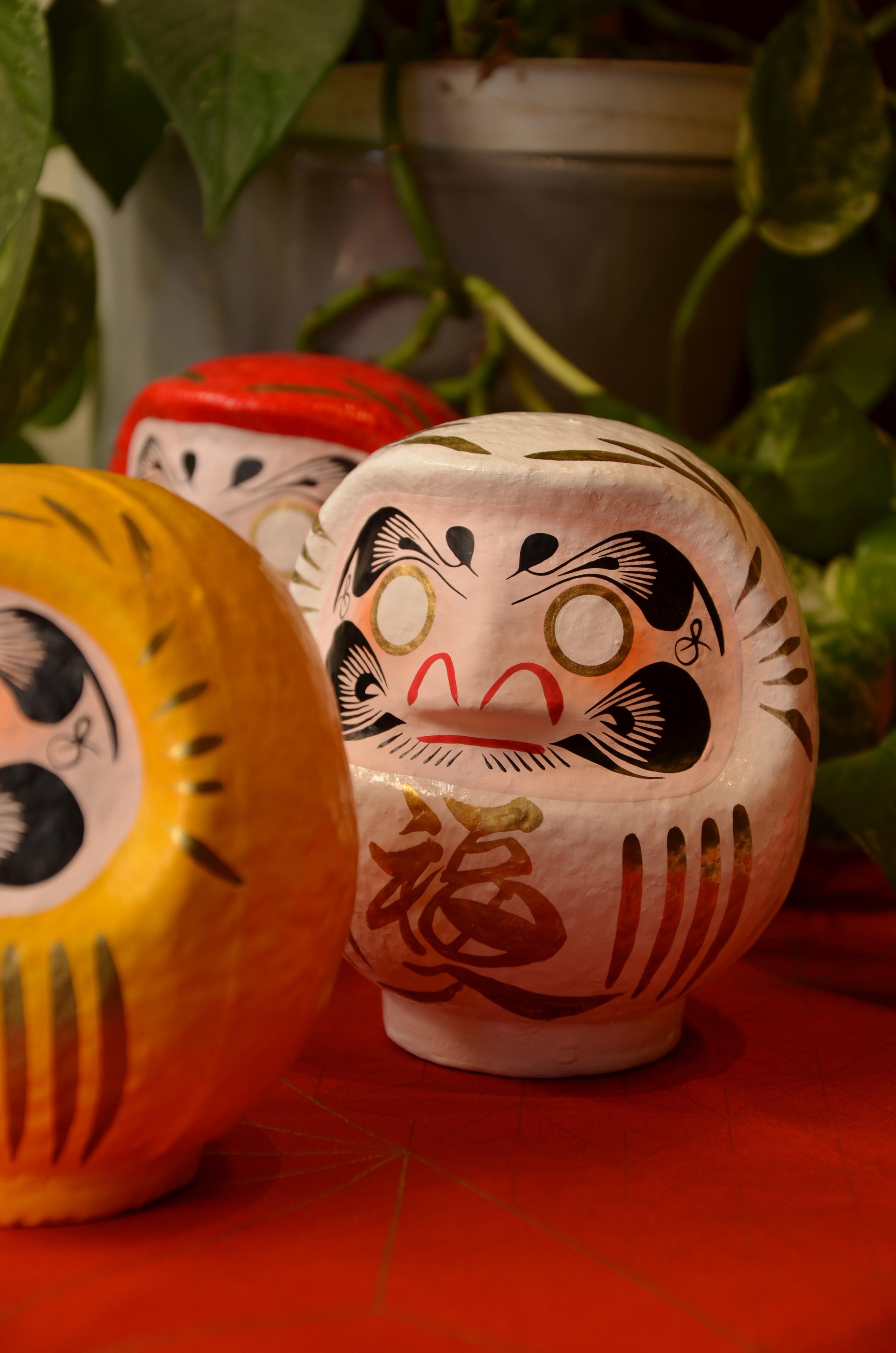 A close-up of a traditional Japanese Daruma doll, painted in white with intricate black, gold, and red details. The doll features a bold, expressive face with large, stylized eyes, a red nose, and a mustache, along with a gold-rimmed circular design on its forehead. A red Japanese kanji character, often symbolizing good fortune or perseverance, is painted on its side. The doll is placed on a red surface, with other colorful Daruma dolls and green foliage softly blurred in the background, creating a vibrant and culturally rich scene.