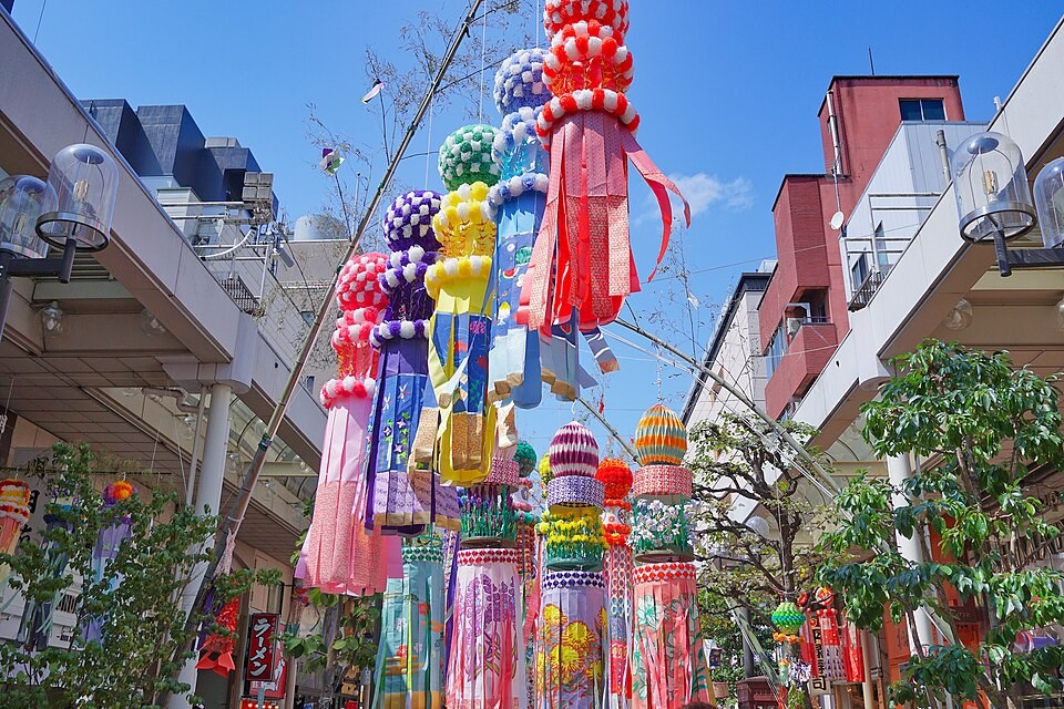 A vibrant and festive scene from the Sendai Tanabata Matsuri, featuring colorful and intricately crafted paper decorations hanging from tall bamboo poles. The decorations include large, round pom-poms in shades of pink, yellow, and purple, along with long, flowing streamers in red, blue, and other bright colors. The bamboo poles are adorned with origami cranes, paper lanterns, and other traditional ornaments, creating a lively and celebratory atmosphere. The decorations are set against a backdrop of modern buildings and a clear blue sky, capturing the blend of tradition and urban life during this iconic Japanese festival.