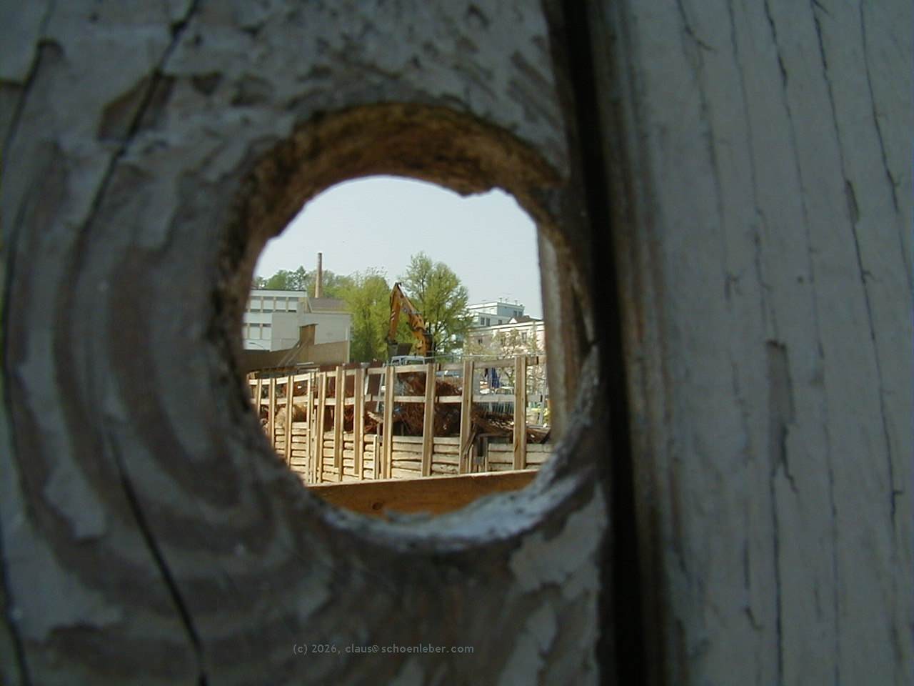 Das Bild zeigt ein Loch in einem Bauzaun (im Vordergrund, dunkel). Der Bauzaun füllt das ganze Bild aus. Durch das Loch sieht man eine Großbaustelle, es ist ein heller Bauzaun zu sehen, der eine Baustelle umrahmt, dahinter ist der Greifarm eines Baggers zu sehen. Ganz im Hintergrund des Bildes, das man durch das kreisfürmige Loch sieht, ist der Gebäudekomplex der Löwenbrauerei in Passau zu sehen. Dazwischen sind einige Laubbäume mit grünen Blättern im Hintergrund zu sehen. Die Baustelle gehörte zum Bau der so genannten "Neuen Mitte", die in Passau die Innenstadt vor einigen Jahren komplett umgestaltete. Es war ein "Otto"-Projekt...